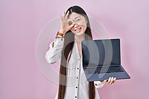 Chinese young woman holding laptop showing screen smiling happy doing ok sign with hand on eye looking through fingers