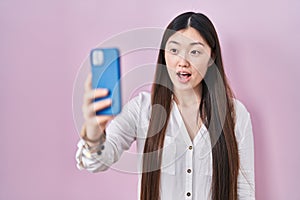 Chinese young woman taking a selfie photo with smartphone scared and amazed with open mouth for surprise, disbelief face