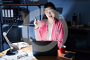 Chinese young woman working at the office at night with a big smile on face, pointing with hand and finger to the side looking at