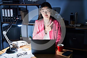 Chinese young woman working at the office at night cheerful with a smile of face pointing with hand and finger up to the side with