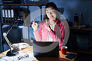 Chinese young woman working at the office at night smiling and looking at the camera pointing with two hands and fingers to the