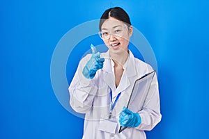 Chinese young woman working at scientist laboratory pointing fingers to camera with happy and funny face