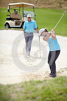 A chip off the old block. A young female golfer chipping her ball out of a bunker while her male partner looks on from