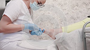 Chiropodist in blue medical mask and gloves gives a pedicure to the client's foot, trimming dry cuticle skin with