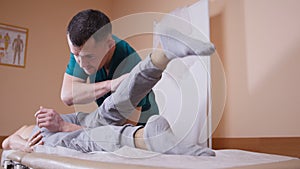 Chiropractor massaging a young woman lying on a massage table, stretching and flexing her hip joint pressing on her knee