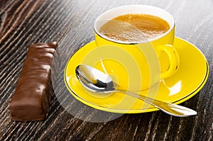 Chocolate bar with filling, cup with black coffee, spoon on saucer on wooden table