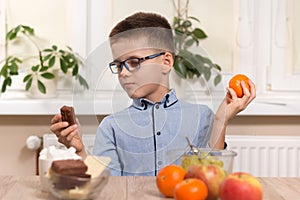 Chocolate bar or tangerine fruit. School-age boy sits at the table and wonders what to choose.