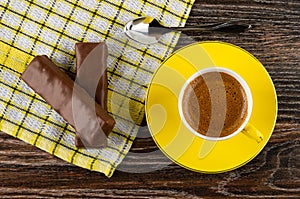 Chocolate bars with filling, spoon on napkin, yellow cup with coffee on saucer on wooden table. Top view