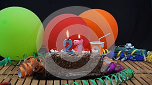 Chocolate birthday 27 cake with candles burning on rustic wooden table with background of colorful balloons