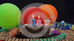 Chocolate birthday 30 cake with candles burning on rustic wooden table with background of colorful balloons