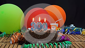 Chocolate birthday 300 cake with candles burning on rustic wooden table with background of colorful balloons