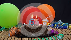 Chocolate birthday 65 cake with candles burning on rustic wooden table with background of colorful balloons