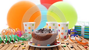 Chocolate birthday cake on rustic wooden table with background of colorful balloons, gifts, plastic cups with candies