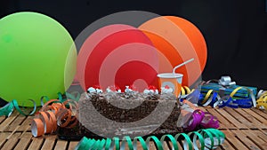 Chocolate birthday cake on rustic wooden table with background of colorful balloons, gifts, plastic cups and streamers