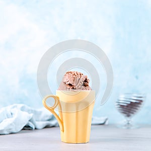 Chocolate brown ice cream in a regular yellow mug, on a blue background. Unusual serve of dessert, copy space