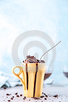 Chocolate brown ice cream in a regular yellow mug and spoon, on a blue background, with topping and crumbs. Unusual serve of