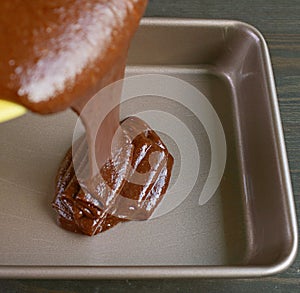 Chocolate Cake Batter Being Poured into Greased Cake Pan with Blurry Cake Mixture in Foreground