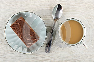 Chocolate cake in blue saucer, spoon, coffee with milk in cup on wooden table. Top view