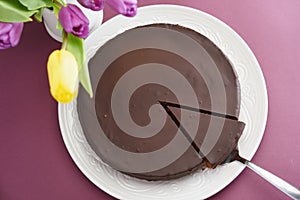 Chocolate cake on a purple table and a bouquet of puprle and yellow tulips. Top view