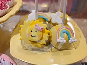 Chocolate cake, small, covered in yellow chocolate decorated with a smiling sun face and with rainbow served on a stick.