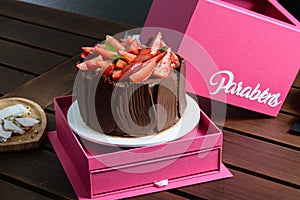 Chocolate cake with strawberries, basil leaves and chocolate plates around, on a pink box. Written in Portuguese: Congratulations. photo