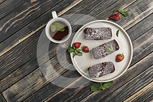 Chocolate cakes with strawberry filling on a flat plate with a cup of tea on a white wooden background