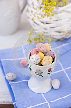 Chocolate candy colored Easter eggs in ceramic cup on blue checkered napkin, basket with flowers