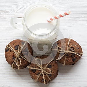 Chocolate chip cookies and glass jar of milk on a white wooden table, overhead view.