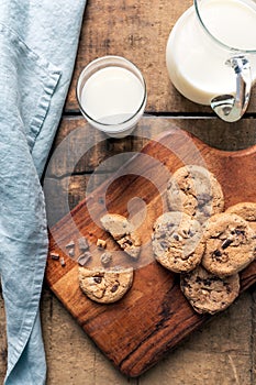 Chocolate chip cookies on wooden cutting board and rustic background with milk mug and milk glass.