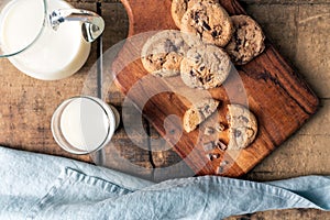 Chocolate chip cookies on wooden cutting board and rustic background with milk mug and milk glass.