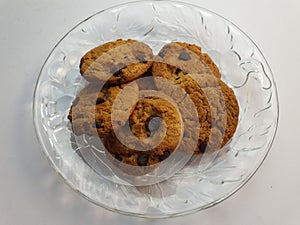 Chocolate chips cookies in a clear round shaped glassware dish plate laid on white surface background.