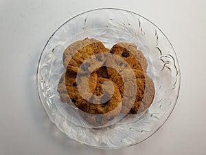 Chocolate chips cookies in a clear round shaped glassware dish plate laid on white surface background.