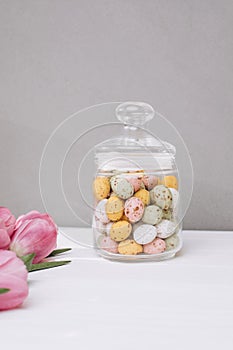 Chocolate colorful decorative eggs in a glass jar and pink tulips on a white wooden table. Happy Easter