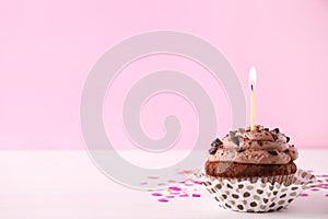 Chocolate cupcake with burning candle on white table against pink background. Space for text