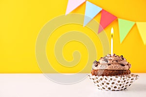 Chocolate cupcake with burning candle on white table against yellow background. Space for text