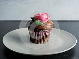 Chocolate cupcake with chocolate frosting topped with a pink frosting rose with green leaves on a plate with a gray and black