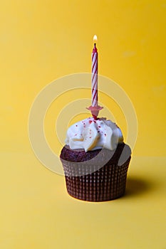 Chocolate cupcake with white cream and a burning candle on a yellow background