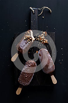 Chocolate dipped popsicles with chipped nuts on dark wooden board over black background