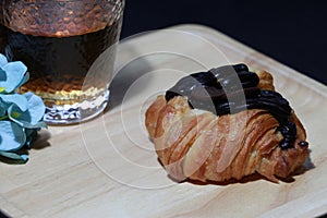Chocolate flavor croissant placed on the square wooden tray with tea in a glass and blue flowers on dark background