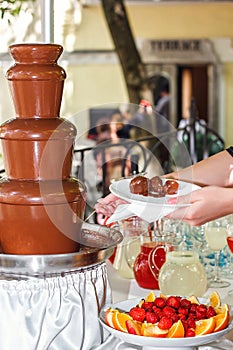 Chocolate fondue with fruits assortment. Female hand dipping strawberry on a skewer into the warm chocolate fondue fountain at the