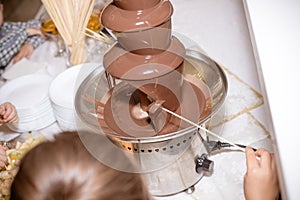 Chocolate Fountain on kids birthday party with a kids playing around and marshmallows and fruits dip dipping into