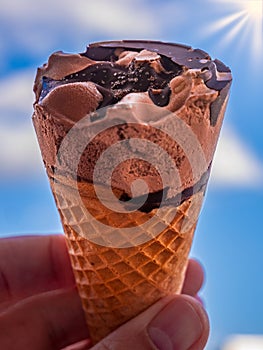 Chocolate ice cream cone held against a summer sky with clouds and sun with rays in the upper right corner