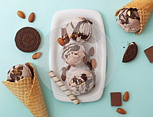Chocolate ice cream in a waffle cone and on a white plate, decorated with crispy cookies, nuts, chocolate pieces, blue