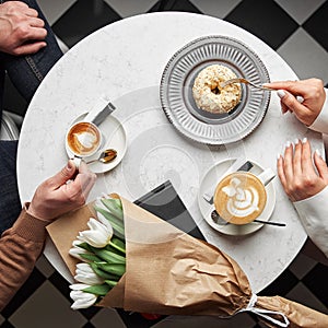 Hands of young couple eating dessert and holding hands on table with coffee, gift box and flowers