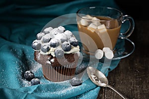 Chocolate muffin with soft cream and fresh blueberry powdered with sugar, cup of coffee with marshmallows, rustic background