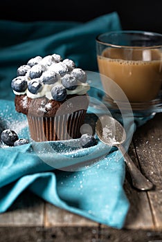 Chocolate muffin with soft cream and fresh blueberry powdered with sugar, cup of coffee, rustic background