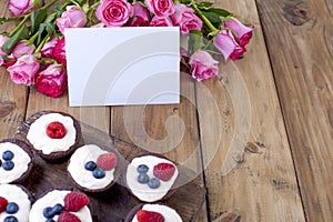 Chocolate muffins with I raspberries and blueberries, and white cheese cream, on a brown wooden board. A bouquet of fresh pink