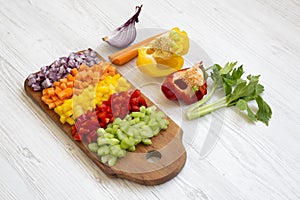 Chopped fresh vegetables arranged on cutting board on white wooden surface, side view.
