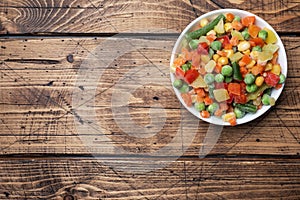 Chopped frozen vegetables in a plate on a wooden background. Corn peas pepper carrots. copy space