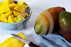 Chopped mango in white bowl and mango with peel on the side and seed on plate with knife